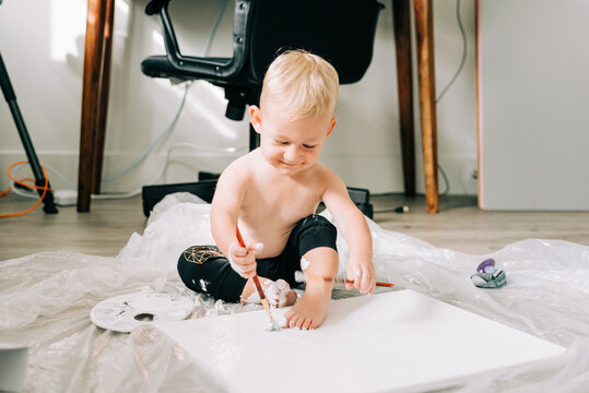 A blonde two year old boy paints on a canvas