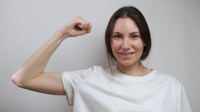 A pretty young adult Caucasian millennial woman smiles, looks at the camera, raises her arm and shows her biceps. The concept of a healthy body