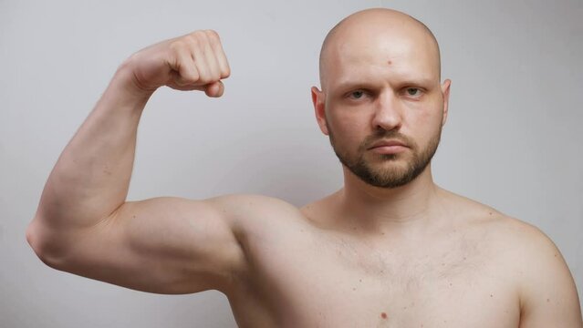 A muscular bald male athlete bodybuilder with a beard and a bare torso looks seriously at the camera, raises his arm, strains and shows his biceps. Sports health, strong physical body