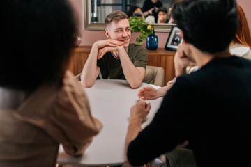 Front view of handsome young man sitting around table with multiethnic friends, telling story, hang out with men and women enjoying free time. Four friends have fun at home, chatting, telling stories.