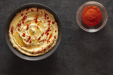 Hummus with olive oil and red pepper in grey bowl, on slate stone plate round, dark background, top view