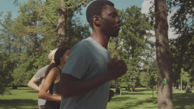 Medium shot of three diverse athletic friends jogging together along track in public park on sunny day