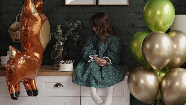 Child birthday party. Adorable happy little girl in festive stylish dress sit on commode on green brick wall background at room decorated with balloons,deer form balloon and birthday cake with candle.