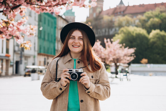 Young beautiful smiling hipster woman in trendy clothes with camera. Carefree woman with backpack posing on the street background. Positive tourist outdoors. Cheerful 30s girl in hat is exploring city