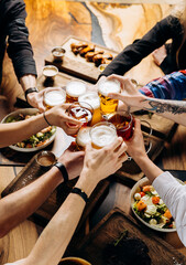 Friends cheering beer glasses on wooden table covered with delicious food - Top view of people...