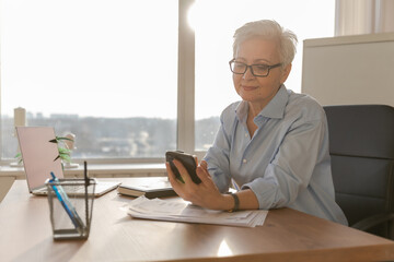 Confident stylish european middle aged senior woman using smartphone at workplace. Stylish older...