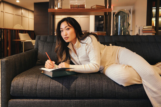 Woman writing with a pencil in a notebook lying on the couch