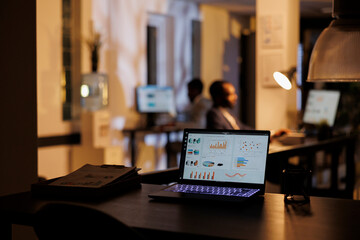 Selective focus on laptop computer with financial graph on screen, standing on desk table in startup office. In background employees working at marketing strategy late at night. Business concept