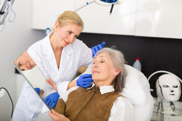 Obraz premium Cosmetologist and her patient, mature woman, looking in mirror after face lifting procedures.