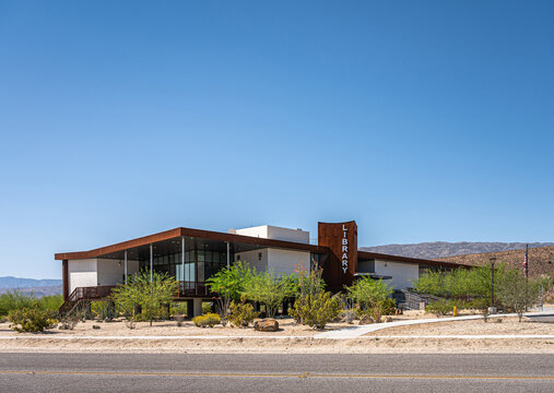 Borrego Springs, CA, USA - April 24, 2023: full frontal view on  modern architecture style public Library building under blue sky, set on sandy desert floor with green bushes. US flag present