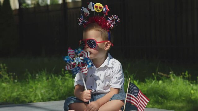funny little kid in red, white, blue accessories sit green grass blow american colors windmill. national American flag is near child, his family celebrate 4 July Independence day, have picnic lawn