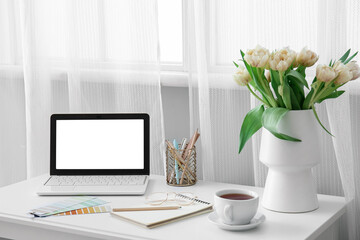 Comfortable workplace with modern laptop, cup of tea and beautiful tulip flowers near window