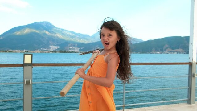 Funny child with a tool. A view of playful happy little girl hold a pickaxe on the wooden pier against blue sea water during summer time.