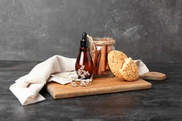 Wooden board with bottle of essential oil, cinnamon sticks and cookies on dark background