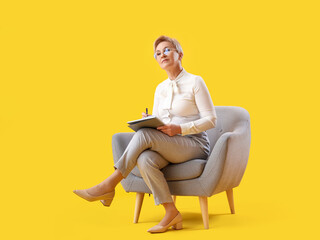 Mature psychologist with notebook sitting in armchair on yellow background - obrazy, fototapety, plakaty