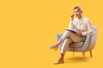 Fototapeta na wymiar Mature psychologist with notebook sitting in armchair on yellow background