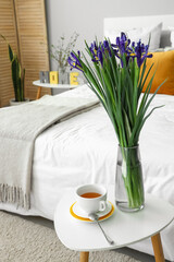 Vase with beautiful iris flowers and cup of tea on bedside table