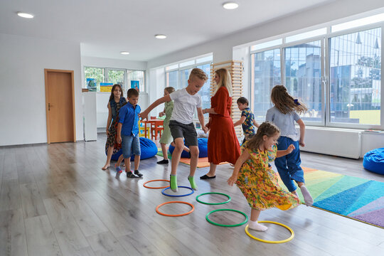 Small nursery school children with female teacher on floor indoors in classroom, doing exercise. Jumping over hula hoop circles track on the floor.
