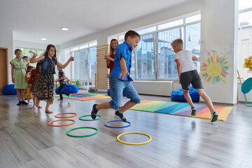 Small nursery school children with female teacher on floor indoors in classroom, doing exercise. Jumping over hula hoop circles track on the floor.