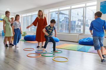 Small nursery school children with female teacher on floor indoors in classroom, doing exercise. Jumping over hula hoop circles track on the floor.