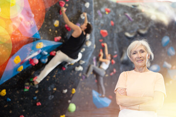 elderly woman posing with her arms crossed over her chest after climbing class. Artificial black bouldering wall