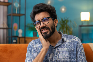 Dental problems. Young indian man touching cheek, closing eyes with expression of terrible suffer from painful toothache, sensitive teeth, cavities. Hindu guy at home apartment living room on sofa