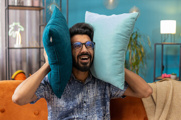 Repair work at neighbours. Irritated indian man relaxing on couch cover ears with pillows annoyed...