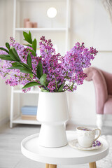 Vase with blooming lilac flowers and cup of tea on coffee table in interior of living room