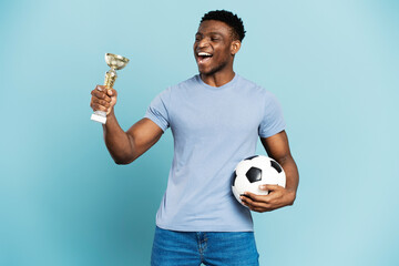 Excited african american football player holding cup and soccer ball, isolated on blue background