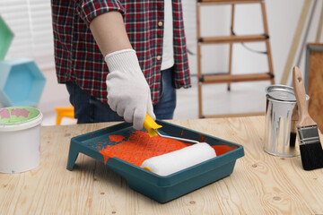 Man taking orange paint with roller from tray at wooden table indoors, closeup