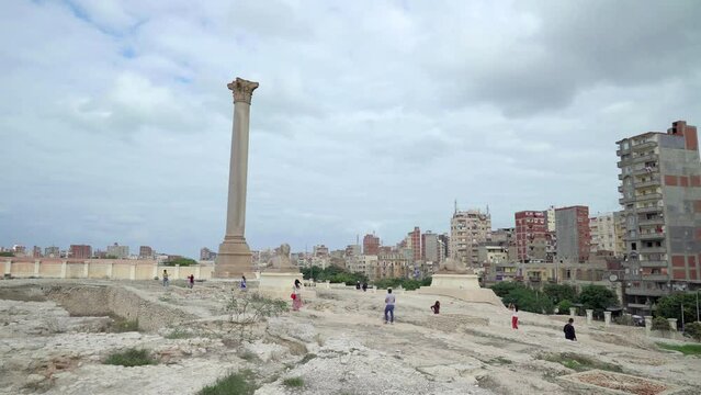 Alexandria, Egypt. December 2022. Panoramic view of the ruins of Serapeum temple. In the background countless buildings. Some tourists take pictures next to Pompey's column and two sphinxes.