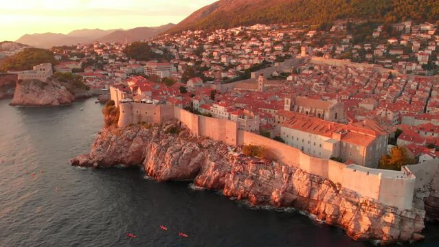 An aerial view of a red sunset over Dubrovnik. People can be seen enjoying the sunset on kayaks in front of the city walls. A castle is visible on the horizon