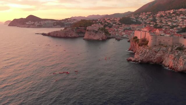 An aerial view of a sunset over Dubrovnik. People can be seen enjoying the sunset on kayaks in front of the city walls. A castle is visible on the horizon