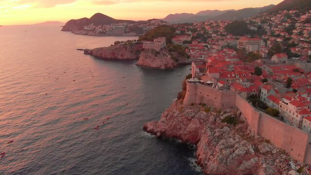 An aerial view of a sunset over Dubrovnik. People can be seen enjoying the sunset on kayaks in front of the city walls. The walls of the old town visible