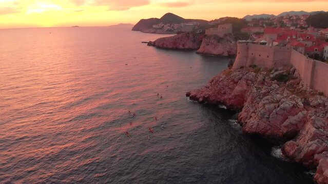 An aerial view of a sunset over Dubrovnik. People can be seen enjoying the sunset on kayaks in front of the city walls. The walls of the old town are visible