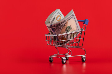 100 american dollars bills in a miniature shopping cart from a supermarket on a red background