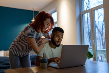 Loving caucasian woman talking with boyfriend working on laptop late at home, smiling girlfriend demanding attention from busy with work African American guy freelancer. Work life balance