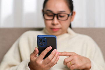Happy young woman using mobile phone while sitting on sofa at home. Attractive woman holding smartphone checking social media, playing games, online shopping relaxing on sofa.