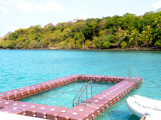 Floating swimming pool made by floating modular plastic blocks with ladder on the tropical blue sea near the speed boat and green hill island on sunny day. Summer pool with ocean view background.