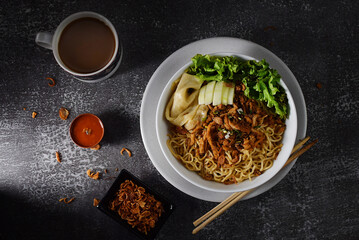 Mie ayam, noodles with chicken and vegetables in white bowl, Indonesian traditional food in dark and texture background. served with fried onions and a cup of coffee. hand holding chopsticks
