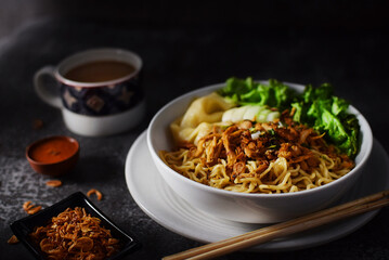 Mie ayam, noodles with chicken and vegetables in white bowl, Indonesian traditional food in dark and texture background. served with fried onions and a cup of coffee