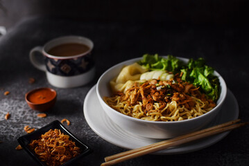 Mie ayam, noodles with chicken and vegetables in white bowl, Indonesian traditional food in dark and texture background. served with a cup of coffee, sauce and fried onions