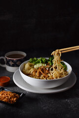 Mie ayam, noodles with chicken and vegetables in white bowl, Indonesian traditional food in dark and texture background. served with fried onions and a cup of coffee