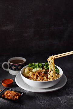 Mie ayam, noodles with chicken and vegetables in white bowl, Indonesian traditional food in dark and texture background. served with fried onions and a cup of coffee. hand holding chopsticks