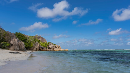 Fototapeta na wymiar A beautiful beach on a tropical island. Picturesque cliffs with steep smoothed slopes and boulders at the water's edge. Turquoise ocean, white sand, blue sky with clouds. Seychelles. La Digue. 