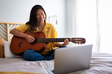 Happy teenage asian girl learning to play guitar with online teacher using laptop in bedroom sitting on bed. Copy space.