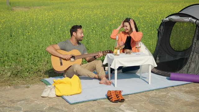 Young Couple in Bright Clothes Sitting By Rapeseed Flowering Meadow Having a Camping Picnic - Man Playing Guitar and Flirting with Prety Woman, Happy Together