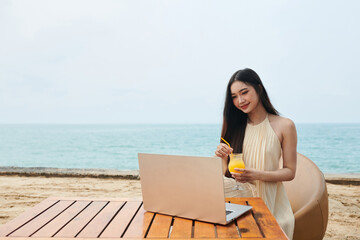 Young woman sitting at table on beach, drinking cocktail and working on laptop