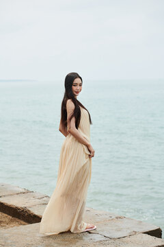 Young Asian woman in long flowing dress standing on beach and smiling at camera