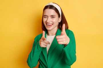 Flirting positive woman wearing green jacket posing isolated over yellow background showing finger guns to camera winking expressing happiness.